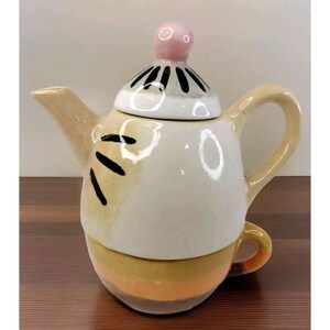 May include: A ceramic teapot and cup set with a whimsical design. The teapot has a rounded body with a yellow, orange, and white striped pattern. The lid has a black and white striped pattern with a pink knob. The cup has a yellow and orange striped pattern.