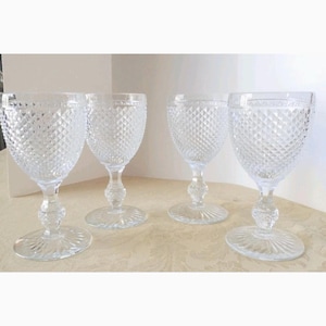 May include: Four clear wine glasses with a diamond pattern, rounded bowl, and decorative stem. The glasses have a round base. They are displayed on a light-coloured surface.