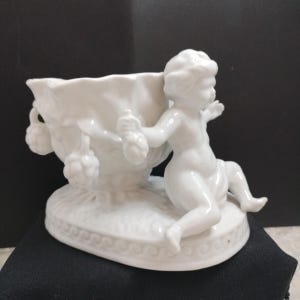 Könnte beinhalten: Eine weiße Porzellanfigur mit einem Putto, der sich an einer dekorativen Schale abstützt. Die Schale ist mit traubenartigen Clustern verziert und ruht auf einem detaillierten Sockel. Die Figur ist ein dekoratives Objekt.