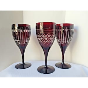 Puede incluir: Tres copas de vino de cristal rojo rubí con intrincados diseños tallados. Las copas tienen un tallo largo y una base ancha y redonda. Los patrones tallados incluyen líneas verticales, círculos y formas de diamante.