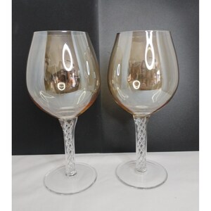 May include: Two wine glasses with iridescent amber-tinted bowls and clear stems featuring a decorative twisted glass lattice pattern. Each glass has a wide, rounded bowl and a circular base, showcasing a reflective surface with light highlights.