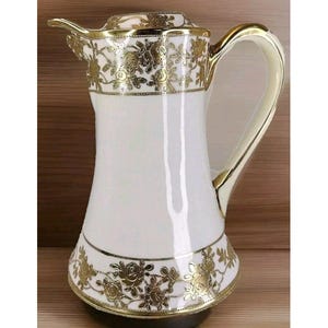 May include: White ceramic pitcher with a gold floral design. The pitcher has a wide base and a narrow neck. The handle is gold and has a slight curve.