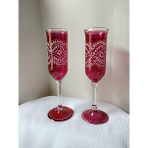 May include: Two ruby red champagne flutes with etched white floral designs. The flutes have a clear stem and base, and the glass is a deep, rich red color. The etched design is a repeating pattern of dots and swirls. The flutes are displayed on a white surface.