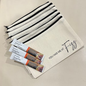 May include: A white canvas zippered pouch with black trim and the text "You had me at Fizz" printed on the front. Three packets of arbonne energy fizz are in front of the pouch.