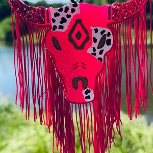 May include: A bright pink bull skull decoration with a black hanging cord. The skull has black and white spotted patterns, a central diamond design, and red fringe. The horns are decorated with glitter.
