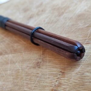 Peut inclure: Un porte-baguettes en bois brun foncé avec une poignée en caoutchouc noir et un anneau noir. Les baguettes ont une pointe arrondie unique. Le support est sur une planche à découper en bois.