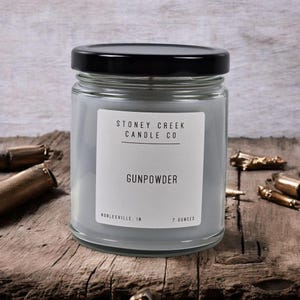 May include: A glass jar candle with a black lid, labeled "Gunpowder" by Stoney Creek Candle Co. The candle is light gray and contains 7 ounces. The background features spent bullet casings on a wooden surface.