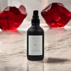 May include: Black room and linen spray bottle with a white label that says "Stoney Creek Candle Co." and "540 Room & Linen Spray". The bottle is sitting on a white marble surface with two red crystal-like objects in the background.