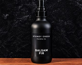 Może przedstawiać: Matowa czarna butelka z rozpylaczem i czarną pompką. Etykieta zawiera biały napis STONEY CREEK Candle Co oraz BALSAM FIR. Butelka ma cylindryczny kształt i gładkie wykończenie, przeznaczona jest do dozowania płynnej zawartości.