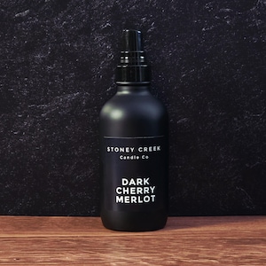 May include: A matte black spray bottle with a black nozzle. The label reads "Stoney Creek Candle Co" and "Dark Cherry Merlot" in white text. The bottle is on a wooden surface.