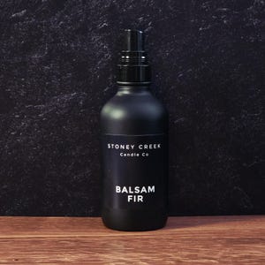 May include: A matte black spray bottle with a black nozzle. The bottle has a white label with the text "STONEY CREEK Candle Co" and "BALSAM FIR". The bottle is on a wooden surface with a dark background.