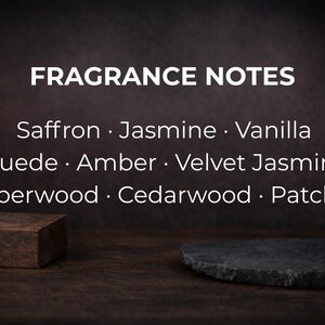 May include: Image featuring the text "FRAGRANCE NOTES" with a list of scent notes: Saffron, Jasmine, Vanilla, Suede, Amber, Velvet Jasmine, Amberwood, Cedarwood, and Patchouli. A small, brown, rectangular block and a dark gray, irregularly shaped stone are in the foreground.