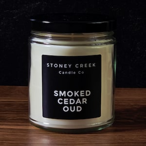 May include: A glass jar candle with a black lid and a black label. The label reads "STONEY CREEK Candle Co" and "SMOKED CEDAR OUD" in white text. The candle has a creamy white wax and sits on a wooden surface.