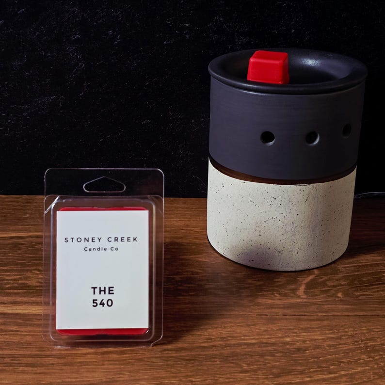 May include: A black and speckled ceramic wax warmer with a red wax cube on top. Next to it is a package of wax melts with the text "Stoney Creek Candle Co" and "THE 540" on the label.