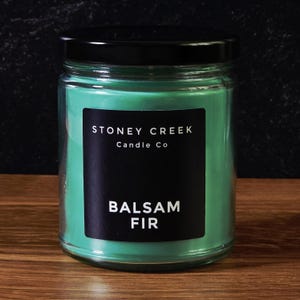 May include: A glass jar candle with a black lid. The candle has a vibrant green wax and a black label that reads "STONEY CREEK Candle Co" and "BALSAM FIR". The candle sits on a wooden surface.