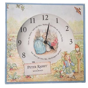 May include: Square clock featuring a Peter Rabbit illustration. The clock face has black hands and a white background with black numerals. The clock is decorated with images of rabbits and garden scenes. The text "PETER RABBIT by WEDGWOOD" is visible.