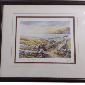 May include: Framed print depicting a coastal scene with a stone wall, path, and figures. The artwork shows a tranquil landscape with water, hills, and a cloudy sky. The frame is dark brown.