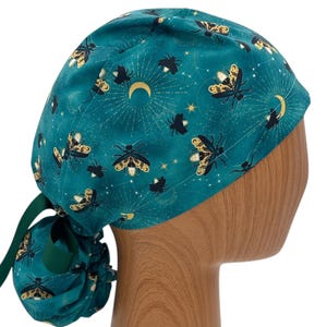 May include: A teal scrub cap featuring a repeating pattern of black and gold fireflies, crescent moons, and small stars. The cap has a gathered back section with a dark green ribbon tie, designed to secure the fabric around the head.