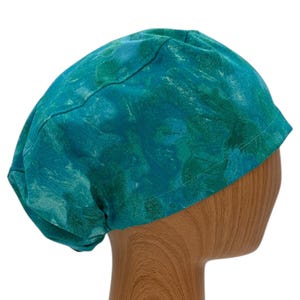 May include: A teal and green patterned scrub cap. The cap is made of fabric and has a rounded shape. The pattern has a watercolor effect with shades of teal and green. The cap is displayed on a wooden mannequin head.