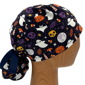 以下が含まれることがあります： 幽霊、カボチャ、キャンディーコーン、星が描かれたハロウィンテーマのプリントが施された黒いスクラブハット。ハットの後ろには青いリボンが付いています。