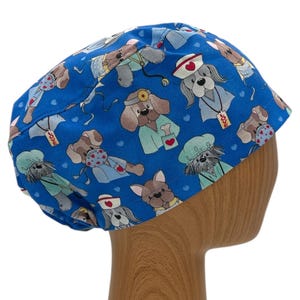 May include: Blue surgical scrub hat with a pattern of cartoon dogs wearing medical scrubs and accessories. The hat has a tie at the back.