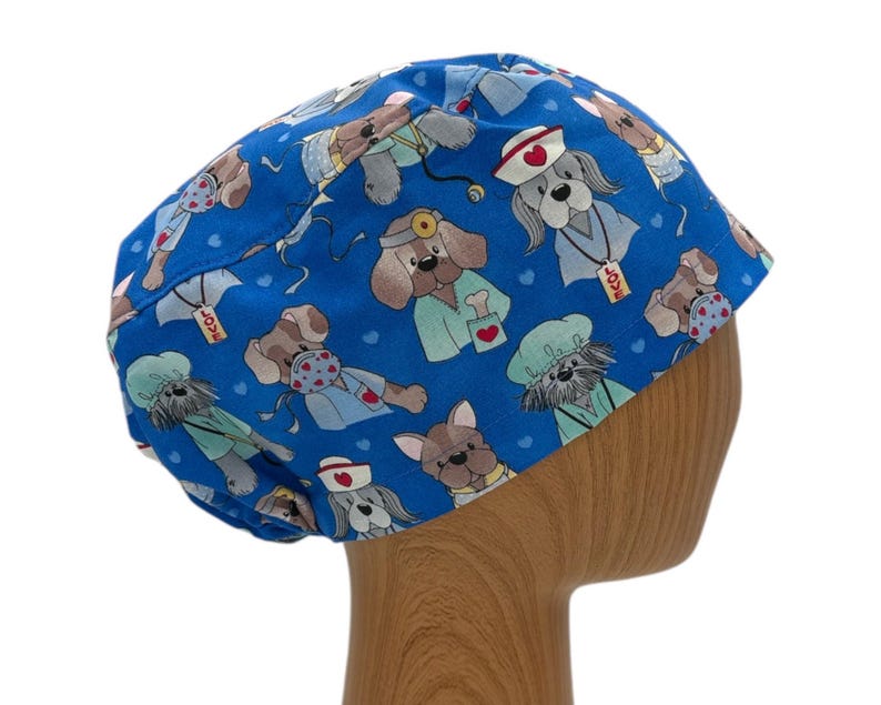 May include: Blue surgical scrub hat with a pattern of cartoon dogs wearing medical scrubs and accessories. The hat has a tie at the back.