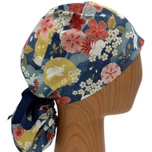 Könnte beinhalten: Eine blaue OP-Mütze mit einem Blumendruck mit weißen Kaninchen und rosa und roten Blumen. Die Mütze hat eine lange Schleife, die am Hinterkopf gebunden werden kann.