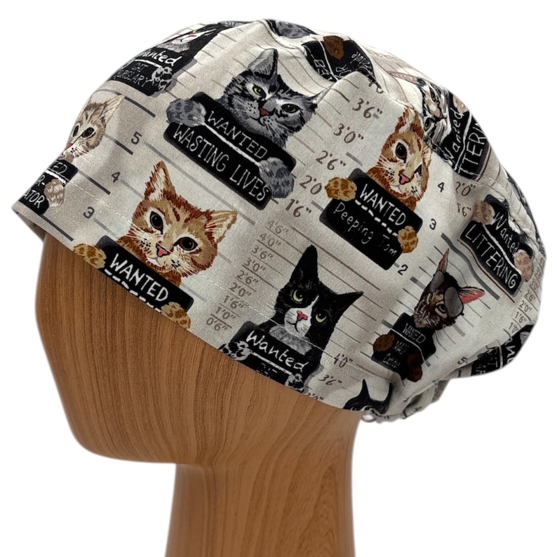 Puede incluir: Un gorro quir&uacute;rgico de color claro con un patr&oacute;n repetido de fotos de gatos. Cada gato sostiene un letrero con frases humor&iacute;sticas como "Wanted: Wasting Lives" y "Wanted: Peeping Tom". El gorro est&aacute; dise&ntilde;ado para adaptarse a la cabeza.
