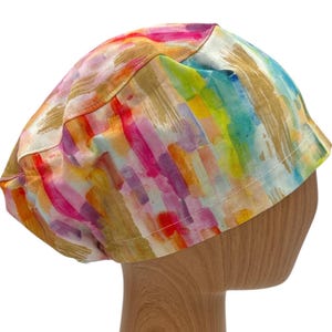May include: A colorful scrub hat with a watercolor-style print featuring shades of pink, purple, blue, green, yellow, and gold. The hat has a white background and a subtle sheen.