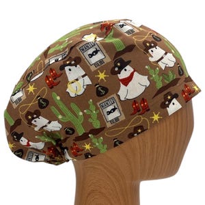 May include: Brown scrub hat with a pattern of white ghosts wearing cowboy hats, cacti, and wanted posters. The text on the posters reads "WANTED $10,000".