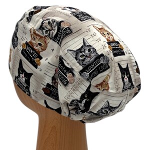 Puede incluir: Un gorro quir&uacute;rgico beige con un patr&oacute;n repetido de gatos que sostienen letreros de "SE BUSCA". Los gatos est&aacute;n representados en varios colores y poses, con texto que dice "Wasting Lives" y "Peeping Toms".