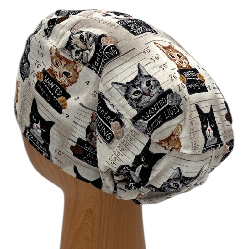 Puede incluir: Un gorro quir&uacute;rgico beige con un patr&oacute;n repetido de gatos que sostienen letreros de "SE BUSCA". Los gatos est&aacute;n representados en varios colores y poses, con texto que dice "Wasting Lives" y "Peeping Toms".