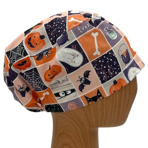 May include: A pink and orange patterned scrub hat with a Halloween theme. The hat features images of pumpkins, bats, ghosts, and a bone with the word "treat" on it.
