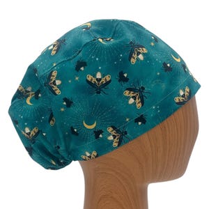 May include: Teal fabric cap with a celestial design featuring moths, moons, stars, and sunbursts. The cap is made of a soft fabric and is designed to fit comfortably. The pattern includes gold and black insect illustrations.