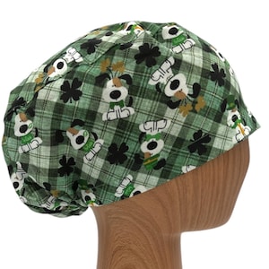 以下が含まれることがあります： 緑と白の格子縞のスクラブハットで、クローバーをつけた漫画の犬の模様が描かれています。犬は白地に黒の斑点があり、茶色の耳と鼻をしています。