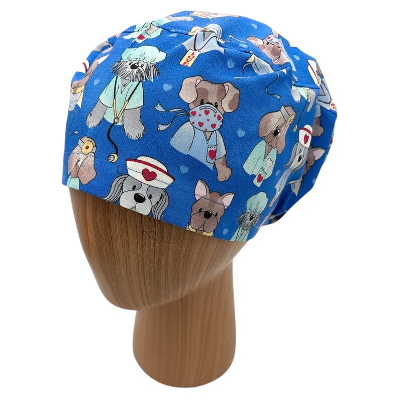 May include: Blue scrub hat with a pattern of cartoon dogs wearing medical masks and scrubs. The dogs are wearing red hearts and some have stethoscopes.