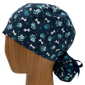 May include: A blue scrub hat with a pattern of paw prints, bones and hearts. The hat has a tie at the back.