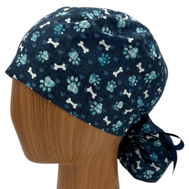 May include: A blue scrub hat with a pattern of paw prints, bones and hearts. The hat has a tie at the back.