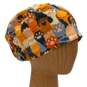 May include: A colorful scrub hat with a pattern of orange and black cat paws and stripes. The hat is on a wooden mannequin head.