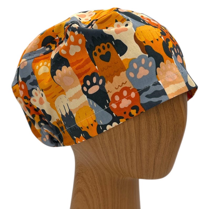 May include: A colorful scrub hat with a pattern of orange and black cat paws and stripes. The hat is on a wooden mannequin head.