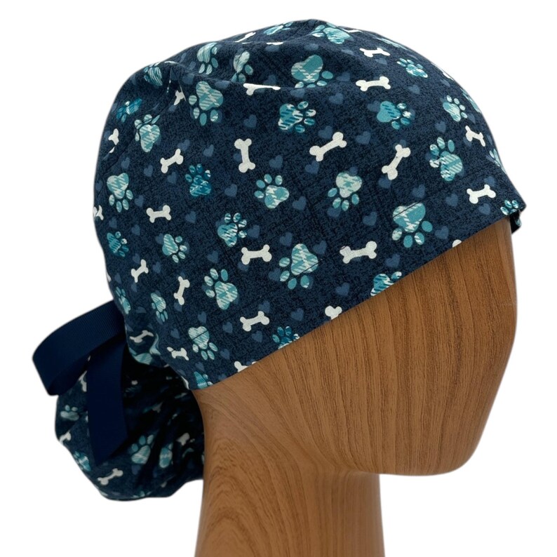 May include: A blue surgical scrub hat with a pattern of paw prints, hearts, and bone shapes. The hat has a navy blue tie.