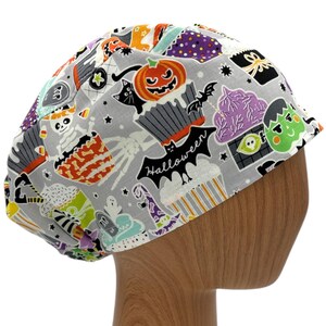 May include: A gray scrub hat with a Halloween themed print featuring cupcakes, bats, ghosts, skeletons, and the word "Halloween".