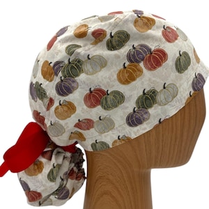 May include: A white scrub hat with a colorful pumpkin print. The hat has a red tie on the back.