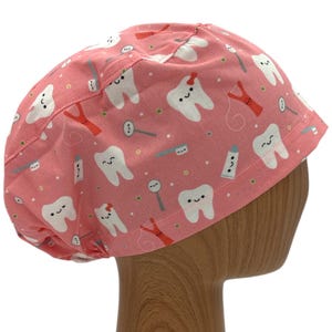 May include: Pink surgical scrub hat with a white tooth pattern. The teeth have smiling faces and are surrounded by dental tools like toothbrushes, floss, and toothpaste.