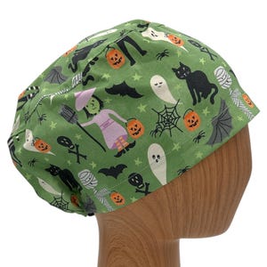 May include: Green scrub hat with a Halloween theme featuring black cats, ghosts, bats, witches, pumpkins, and spiderwebs.