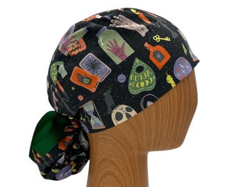 May include: A black scrub hat with a green tie. The hat has a spooky Halloween print with images of hands, bottles, keys, and a Ouija board.