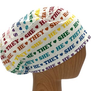 May include: White scrub hat with a rainbow pattern and the words "She, He, They" repeated in a colourful design.