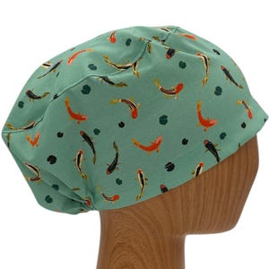 May include: A teal blue scrub hat with a pattern of orange and black fish. The hat is on a wooden mannequin head.