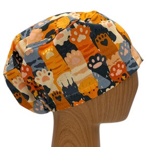 May include: A colorful scrub hat with a pattern of orange, black, and white cat paws. The hat is made of a soft, comfortable fabric and is perfect for nurses, doctors, and other healthcare professionals.