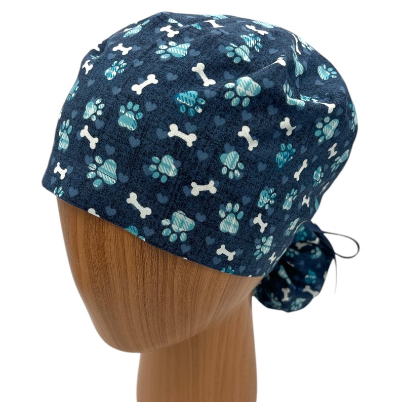 May include: A blue and teal patterned surgical scrub hat with paw prints, hearts, and bone designs. The hat has a tie at the back.